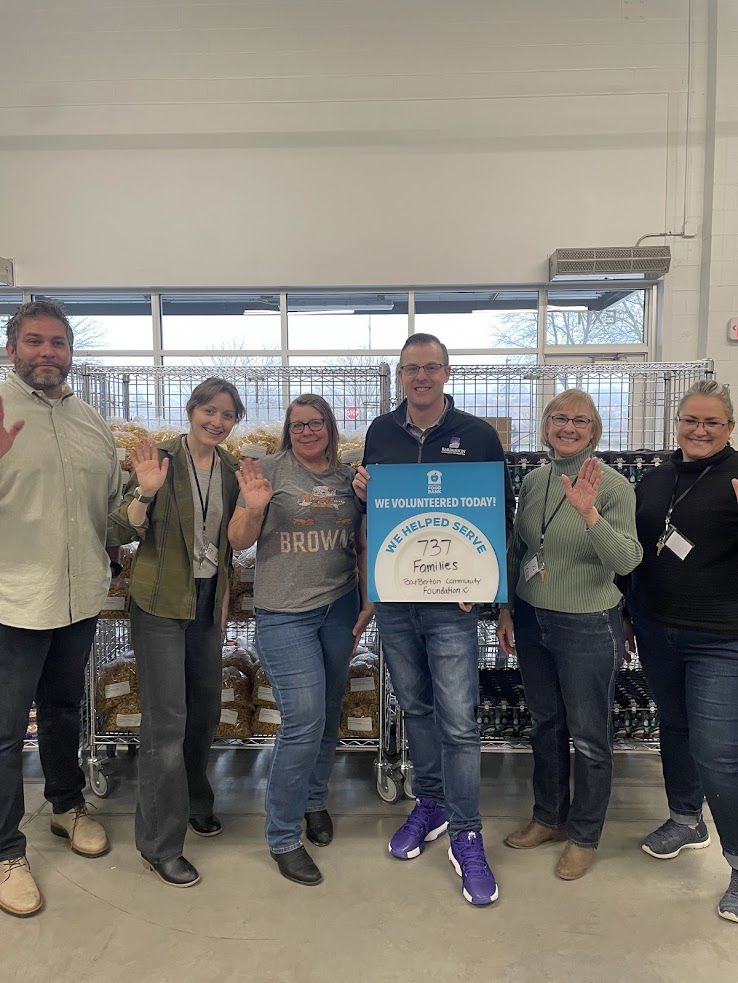 A group of six BCF staff smiling and waving, standing indoors in front of shelves with goods at the Akron Canton Food Bank. They hold a sign reading "We helped serve 737 families today."